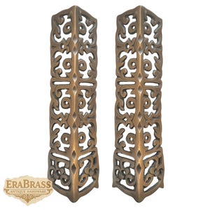 May include: Two antique brass corner brackets with ornate, openwork designs. The brackets are rectangular with a corner shape, featuring scrolling and floral patterns. The EraBrass Antique Hardware logo is visible in the lower left corner.
