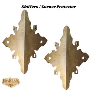 May include: Two brass corner protectors with a decorative, leaf-like design. Each protector has a pointed end and multiple holes for mounting. The text "Skiffers / Corner Protector" is at the top. The logo "EraBrass Antique Hardware" is at the bottom.