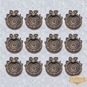 Può includere: Dodici pomelli per mobili antichi in bronzo con un motivo floreale centrale e un fiocco decorativo in alto. I pomelli sono rotondi con una maniglia ad anello e sono disposti a griglia. Il logo EraBrass si trova in basso a destra.