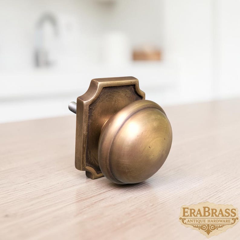 May include: A brass cabinet knob featuring a rounded, dome-shaped handle attached to a square backplate with beveled edges. The metal has a warm, aged finish with visible patina. A threaded metal bolt extends from the back of the hardware piece.