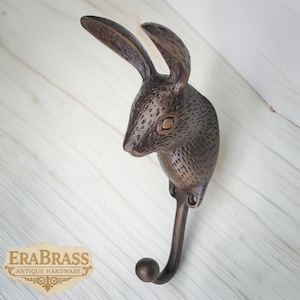 Könnte beinhalten: Ein bronzefarbener Wandhaken in Form eines Kaninchenkopfes. Der Haken ist am unteren Ende des Kaninchenkopfes befestigt und hat ein abgerundetes Ende. Der Haken ist auf einer hellen Holzmaserung montiert. Das Logo "EraBrass Antique Hardware" ist sichtbar.