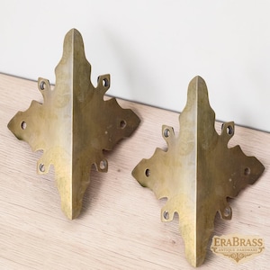 Pode incluir: Dois protetores de canto de latão correspondentes com um formato de diamante pontiagudo e ornamentado, bordas recortadas e quatro orifícios para parafusos pré-perfurados cada. O metal tem um acabamento dourado envelhecido. O texto EraBrass Antique Hardware é visível.