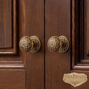 May include: Two round brass cabinet knobs with an intricate star and sunburst pattern, mounted on dark brown wooden cabinet doors. The knobs feature a textured, antique finish with a circular base plate and a central radiating design.