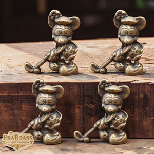 May include: Four antique brass Mickey Mouse figurines. Each figurine is in a seated pose, holding a long, curved object. The figurines are on a wooden surface. The EraBrass logo is visible in the lower left corner.