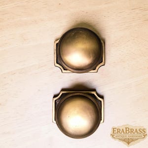 May include: Two circular brass cabinet knobs with a smooth, domed surface and a rectangular backplate featuring decorative notched corners. The metal has a muted, brushed gold finish. The text EraBrass Antique Hardware is visible.