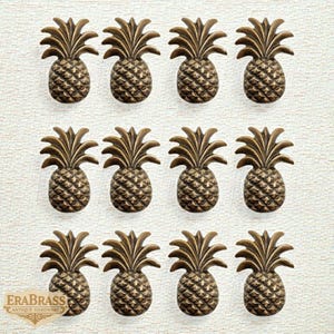 May include: Twelve bronze pineapple-shaped cabinet knobs arranged in three rows of four. Each knob has detailed textures and a classic pineapple design, ideal for adding a tropical touch to furniture. The EraBrass logo is visible in the bottom left corner.