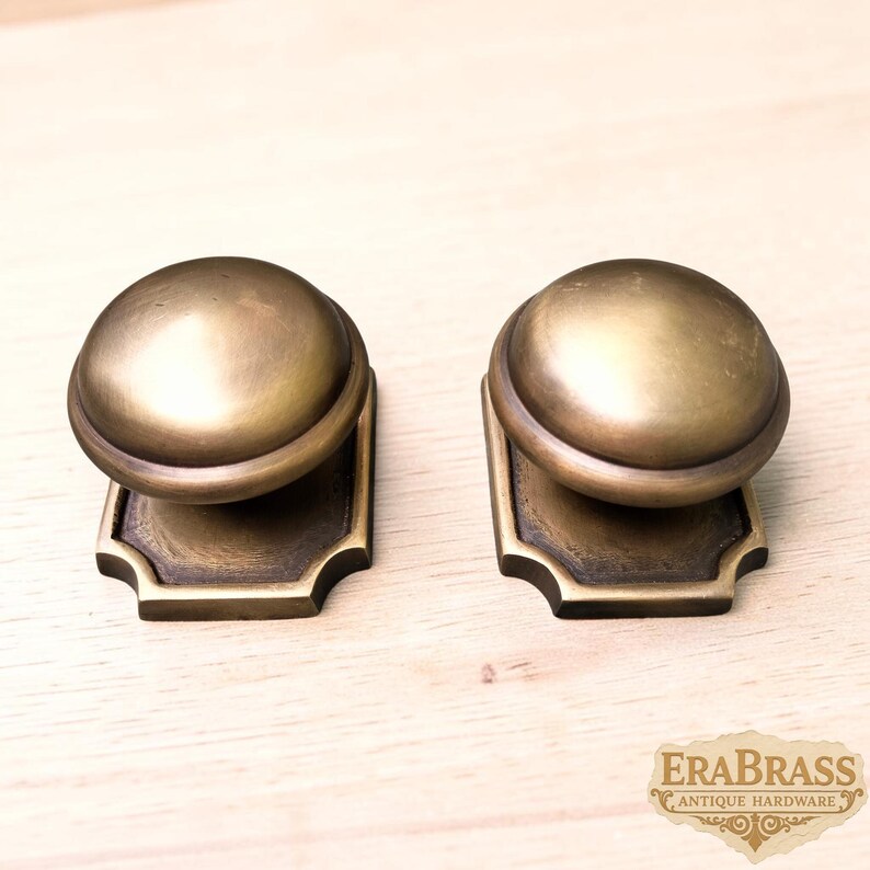 May include: Two matching brass cabinet knobs with a smooth, rounded dome top and a rectangular base plate featuring notched corners. The metal has a muted, aged finish with dark highlights in the recessed areas of the base and the knob stem.