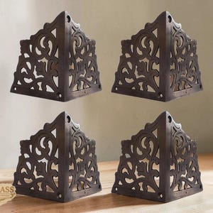 May include: Four antique bronze corner brackets with ornate, openwork designs. Each bracket has a triangular shape with a decorative floral pattern. The brackets are displayed on a wooden surface, showcasing their vintage aesthetic.