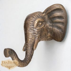 Puede incluir: Un gancho de pared con forma de cabeza de elefante de color bronce. El gancho está formado por la trompa del elefante, que se curva hacia abajo. La cabeza está detallada con arrugas y rasgos, y el gancho está montado sobre una superficie blanca. El logotipo "EraBrass Antique Hardware" es visible.