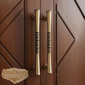 May include: Two brass door pulls with a decorative black and gold band in the center. The pulls are mounted on a dark brown wooden door. The logo "ERABRASS ANTIQUE HARDWARE" is visible in the lower left corner.