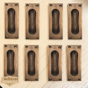 May include: Eight antique brass recessed pulls. Each rectangular pull has a central oval finger pull and four screw holes. The pulls are arranged in two rows of four on a light-colored wooden surface. The logo "ERABRASS ANTIQUE HARDWARE" is visible.