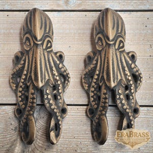 Peut inclure: Deux poignées de meubles en forme de poulpe de couleur bronze. Chaque poignée présente des tentacules détaillées et une surface texturée. Les poignées sont montées sur une surface en bois de couleur claire. Le logo Era Brass est visible en bas à droite.