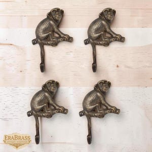 May include: Four antique brass monkey hooks. Each hook features a detailed monkey figure perched on a branch, with a downward-facing hook. The hooks are mounted on a light-colored wooden surface. The logo "ERABRASS ANTIQUE HARDWARE" is visible.