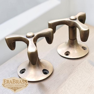 Può includere: Due attaccapanni antichi in ottone con un design a tre punte. Gli attaccapanni hanno una base circolare con tre fori per le viti. Il logo "EraBrass Antique Hardware" è visibile in basso a sinistra.