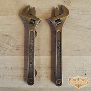 May include: Two brass-colored metal adjustable wrenches shaped as handles. The left wrench is embossed with the text FORGED STEEL, and the right wrench is embossed with 18"-450mm. Both feature a knurled adjustment screw and a circular hole at the base.