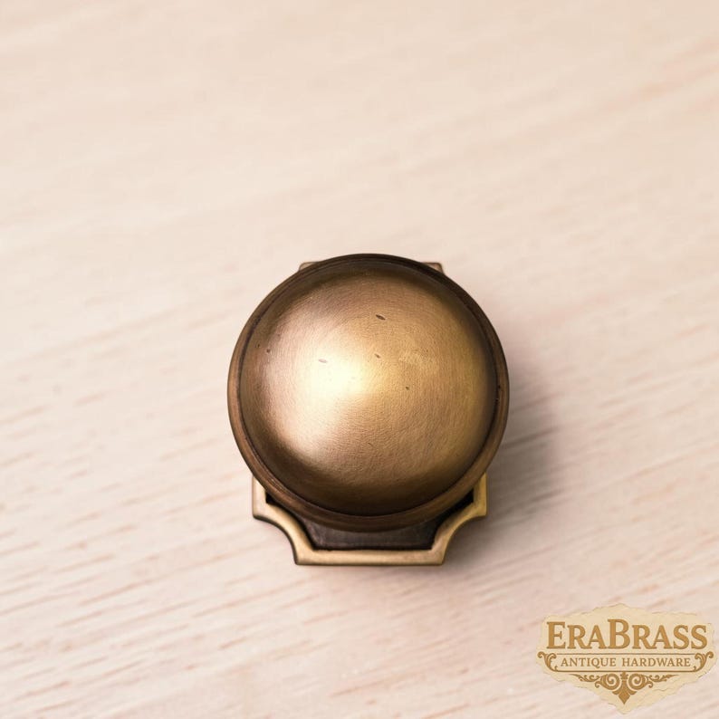 May include: A round, brass-toned cabinet knob with a smooth, domed top and a square base featuring decorative cut-out corners. The metal surface has a matte, aged finish. The text "ERABRASS ANTIQUE HARDWARE" is visible in the bottom right corner.