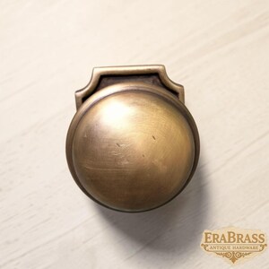 May include: A round brass door knob with a smooth, brushed finish and a rectangular backplate featuring a decorative top edge. The metal surface displays subtle variations in tone and texture, with the text ERABRASS ANTIQUE HARDWARE visible.