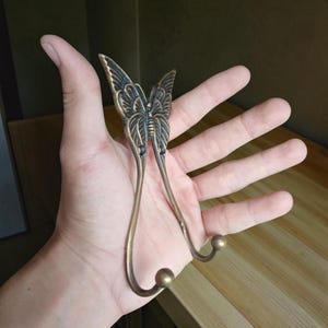 May include: A decorative wall hook with a butterfly design. The hook is made of bronze-colored metal and features detailed wing patterns. The hook has two curved arms with rounded ends for hanging items.