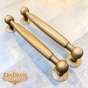 May include: Two antique brass door pulls with a golden finish. Each pull features a cylindrical handle with rounded ends and circular base plates. The EraBrass logo is visible in the lower left corner.