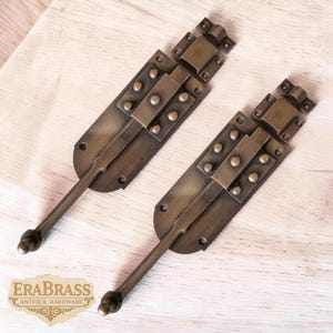 May include: Two antique brass sliding door bolts. Each bolt features a rectangular base with rivets, a sliding bar, and a handle. The bolts are a warm, aged brass color, and the logo "ERABRASS ANTIQUE HARDWARE" is visible.
