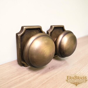 May include: Two round brass door knobs with a brushed antique finish, each mounted on a square backplate with decorative beveled edges. The metal has a warm, muted gold tone with visible texture. The text EraBrass Antique Hardware is visible.