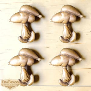 May include: Four antique brass Snoopy cabinet knobs. Each knob features a detailed Snoopy design in a seated position, with a textured, aged finish. The knobs are mounted on a light-colored wooden surface.