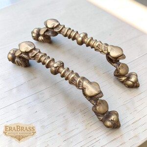 Pode incluir: Duas puxadores de armário antigos de latão. Cada puxador apresenta uma série de desenhos em forma de coração ao longo de uma barra central texturizada. As extremidades dos puxadores têm detalhes decorativos florais ou em forma de folha. Os puxadores são exibidos em uma superfície de madeira clara.