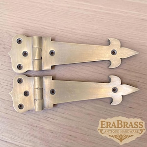 May include: Two matching brass strap hinges with a decorative fleur-de-lis style pointed end and a wide mounting plate featuring three screw holes. The metal has a brushed finish. A logo in the corner reads EraBrass Antique Hardware.