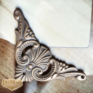 May include: Ornate, bronze-colored corner bracket with a detailed floral and scroll design. The antique hardware piece features a curved shape with multiple mounting holes. The Era Brass Antique Hardware logo is visible in the lower left corner.