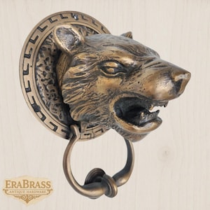 May include: A bronze-colored door knocker in the shape of a snarling bear's head. The knocker has a circular backplate with a decorative border. The handle is a ring attached to the bear's mouth. The words "ERA BRASS ANTIQUE HARDWARE" are at the bottom.
