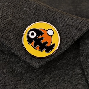 May include: A circular enamel pin featuring a black fish skeleton design on a yellow backdrop, with orange and white accents. The pin is set against a dark grey fabric.