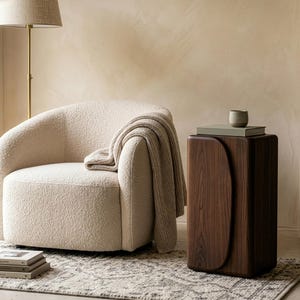 Op de afbeelding: Een gezellige woonkamer met een cr&egrave;mekleurige boucle fauteuil en een gedrapeerde deken. Een donkerhouten bijzettafel met een gebogen ontwerp houdt boeken en een kleine keramische beker vast. Een vloerlamp en een patroonkleed maken de sc&egrave;ne af.