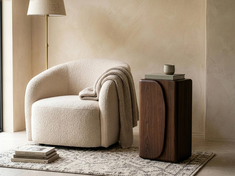 Op de afbeelding: Een gezellige woonkamer met een cr&egrave;mekleurige boucle fauteuil en een gedrapeerde deken. Een donkerhouten bijzettafel met een gebogen ontwerp houdt boeken en een kleine keramische beker vast. Een vloerlamp en een patroonkleed maken de sc&egrave;ne af.