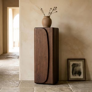 Op de afbeelding: Een hoge, donkere houten kast met een gebogen ontwerp aan de voorkant. Bovenop staat een kleine, bruine vaas met gedroogde stelen. Een ingelijst abstract kunstwerk staat op de vloer naast de kast. Decoratief meubelstuk.