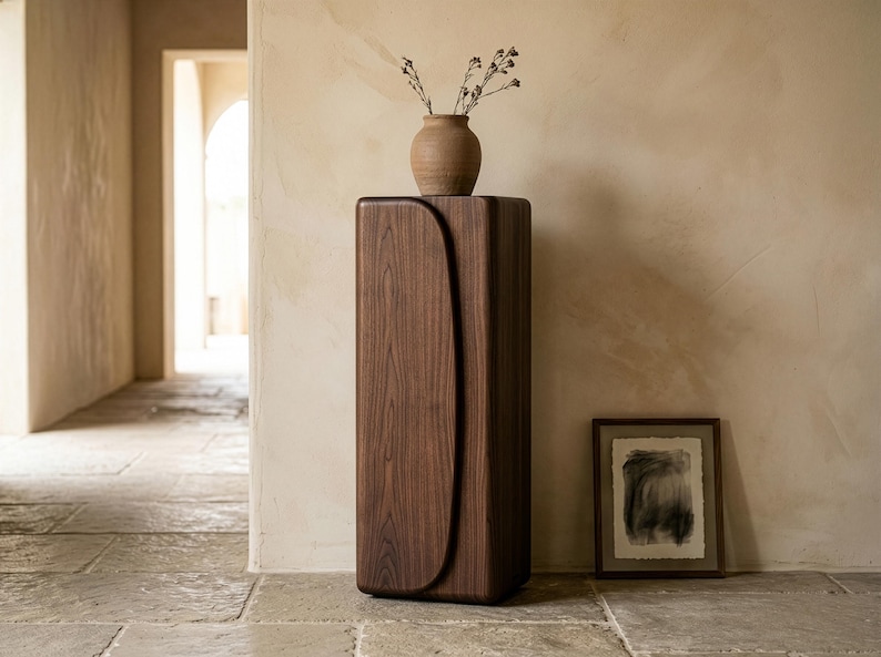 Op de afbeelding: Een hoge, donkere houten kast met een gebogen ontwerp aan de voorkant. Bovenop staat een kleine, bruine vaas met gedroogde stelen. Een ingelijst abstract kunstwerk staat op de vloer naast de kast. Decoratief meubelstuk.