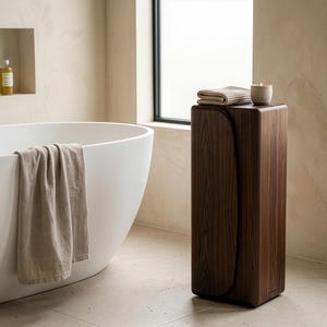Op de afbeelding: Een moderne badkamer met een witte, ovale badkuip en een beige handdoek. Een donkerhouten, rechthoekige bijzettafel met een gevouwen beige handdoek en een kleine kaars. De kamer heeft een neutraal kleurenpalet.