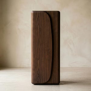 Op de afbeelding: Een hoge, rechthoekige houten kast met een donkerbruine afwerking. De kast heeft een gebogen, lichter gekleurd houten paneel aan de voorkant. Het ontwerp is minimalistisch en modern, met afgeronde randen. De kast wordt waarschijnlijk gebruikt voor opslag.