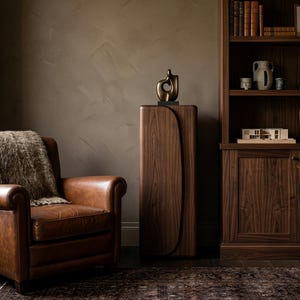 Op de afbeelding: Een gezellige woonkamer met een bruine leren fauteuil en een imitatiebont deken. Een houten kast met een bronzen sculptuur staat in de buurt. Een houten boekenkast en bureau met een lamp maken de ruimte compleet.