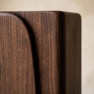 Op de afbeelding: Close-up van een houten hoek van een kast. De kast is gemaakt van donkerbruin hout met een zichtbare nerf. Een gebogen designelement is in het hout gesneden en voegt een vleugje elegantie toe. De achtergrond is neutraal beige.