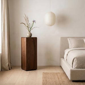 Op de afbeelding: Een moderne slaapkamer met een houten sokkel met een vaas met gedroogde bloemen en een iris. Een papieren lantaarn hangt aan het plafond. Het bed heeft neutrale beddengoed en een geweven vloerkleed.
