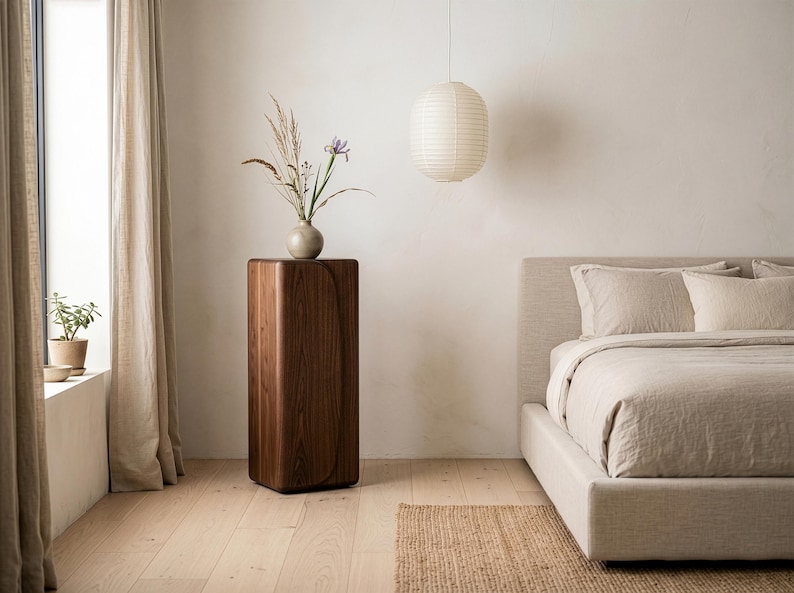 Op de afbeelding: Een moderne slaapkamer met een houten sokkel met een vaas met gedroogde bloemen en een iris. Een papieren lantaarn hangt aan het plafond. Het bed heeft neutrale beddengoed en een geweven vloerkleed.