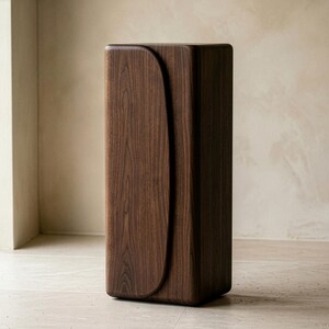 Op de afbeelding: Een hoge, rechthoekige houten kast met een donkerbruine afwerking. De kast heeft een gebogen designelement aan de voorkant. Het meubelstuk staat tegen een neutrale achtergrond en benadrukt de moderne, minimalistische esthetiek.