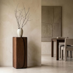 Op de afbeelding: Een modern interieur met een donkerhouten sokkel met een gebogen ontwerp, bekroond met een witte vaas met kale takken. De kamer heeft een neutraal kleurenpalet, met een eettafel en stoelen op de achtergrond.