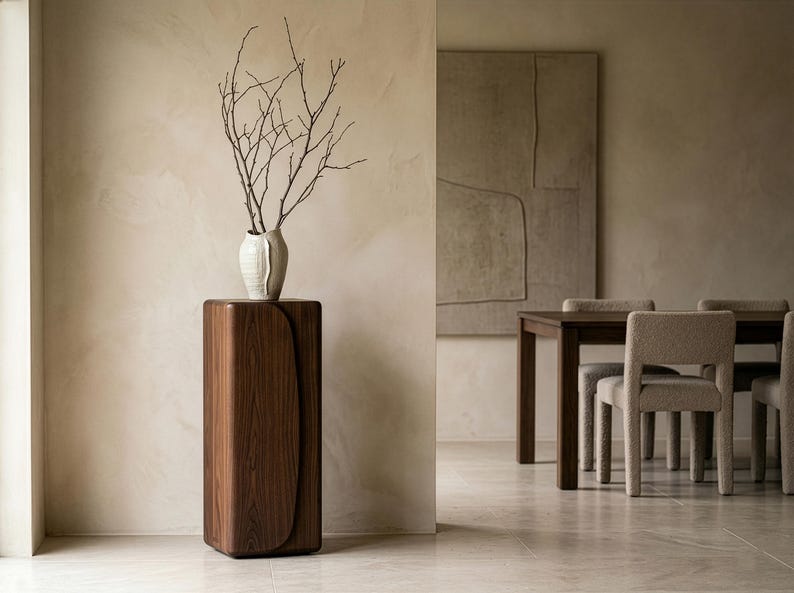Op de afbeelding: Een modern interieur met een donkerhouten sokkel met een gebogen ontwerp, bekroond met een witte vaas met kale takken. De kamer heeft een neutraal kleurenpalet, met een eettafel en stoelen op de achtergrond.