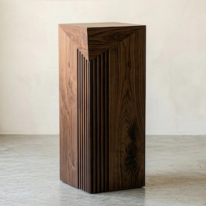 May include: A tall, rectangular wooden pedestal with a dark brown finish. The top features a geometric design, and one side has vertical grooves. The pedestal is a decorative furniture piece.