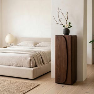 Op de afbeelding: Een moderne slaapkamer met een bed met neutrale beddengoed, een geweven vloerkleed en een houten nachtkastje. Een donkere houten kast met een gebogen ontwerp houdt een vaas met takken en een bloem vast. Een potplant voegt groen toe.