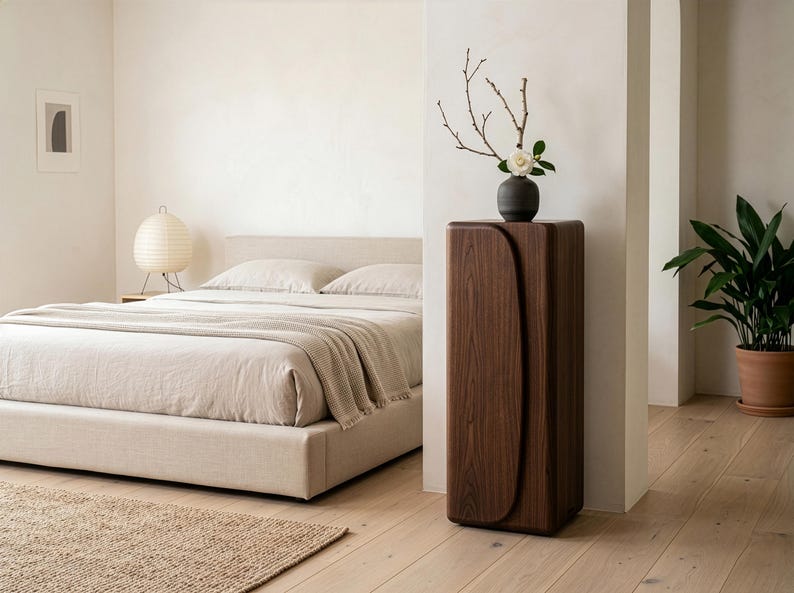 Op de afbeelding: Een moderne slaapkamer met een bed met neutrale beddengoed, een geweven vloerkleed en een houten nachtkastje. Een donkere houten kast met een gebogen ontwerp houdt een vaas met takken en een bloem vast. Een potplant voegt groen toe.