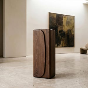 Op de afbeelding: Een hoge, rechthoekige houten kast met een gebogen ontwerp aan de voorkant. De kast is gemaakt van donkerbruin hout en staat op een lichtgekleurde tegelvloer. Op de achtergrond een schilderij en een beige stoel.