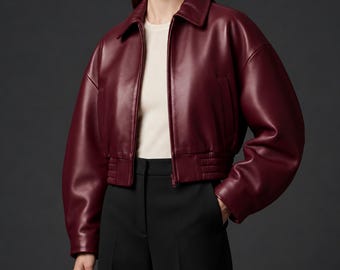 May include: A deep burgundy cropped jacket with a smooth finish, featuring a classic collar, long sleeves with dropped shoulders, a front zipper closure, and a ribbed hemline. The jacket is worn over a plain cream-colored top and black trousers.