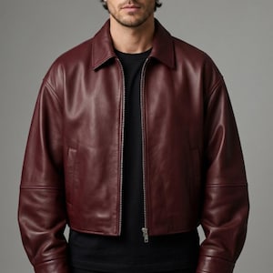 May include: A deep burgundy leather jacket featuring a classic collar, long sleeves with cuffs, and a full-length front zipper closure. The jacket is worn over a plain black t-shirt, showcasing a smooth, matte finish and a structured, waist-length fit.